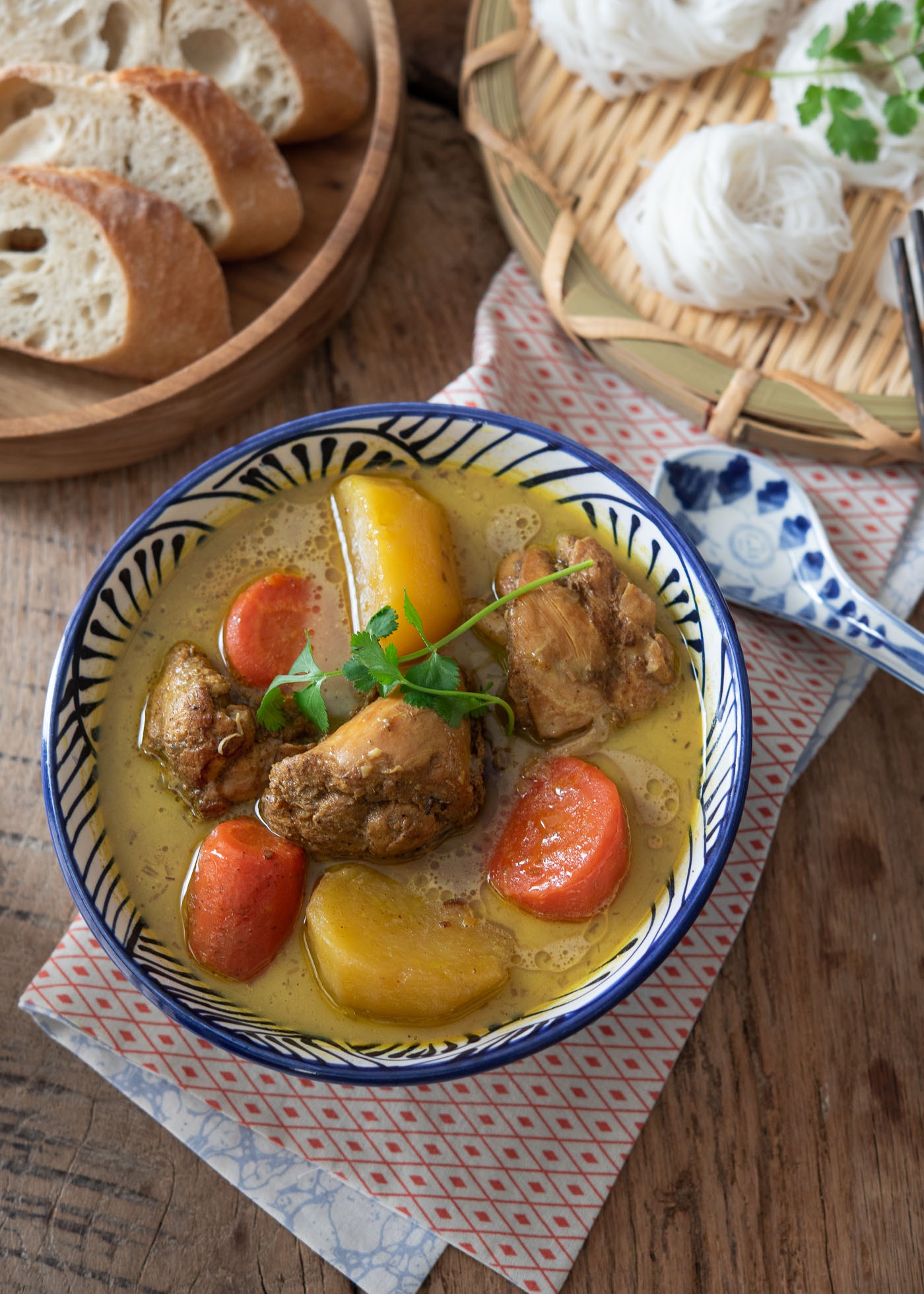 A bowl of chicken curry with potato and carrot are placed on a napkin and paired with baguette slices and rice noodle nests on the side.