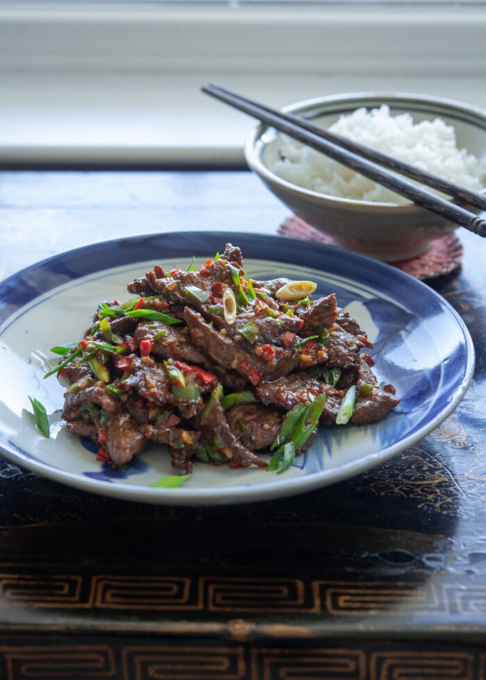 A plate of cumin beef is placed with a bowl of rice and chopsticks