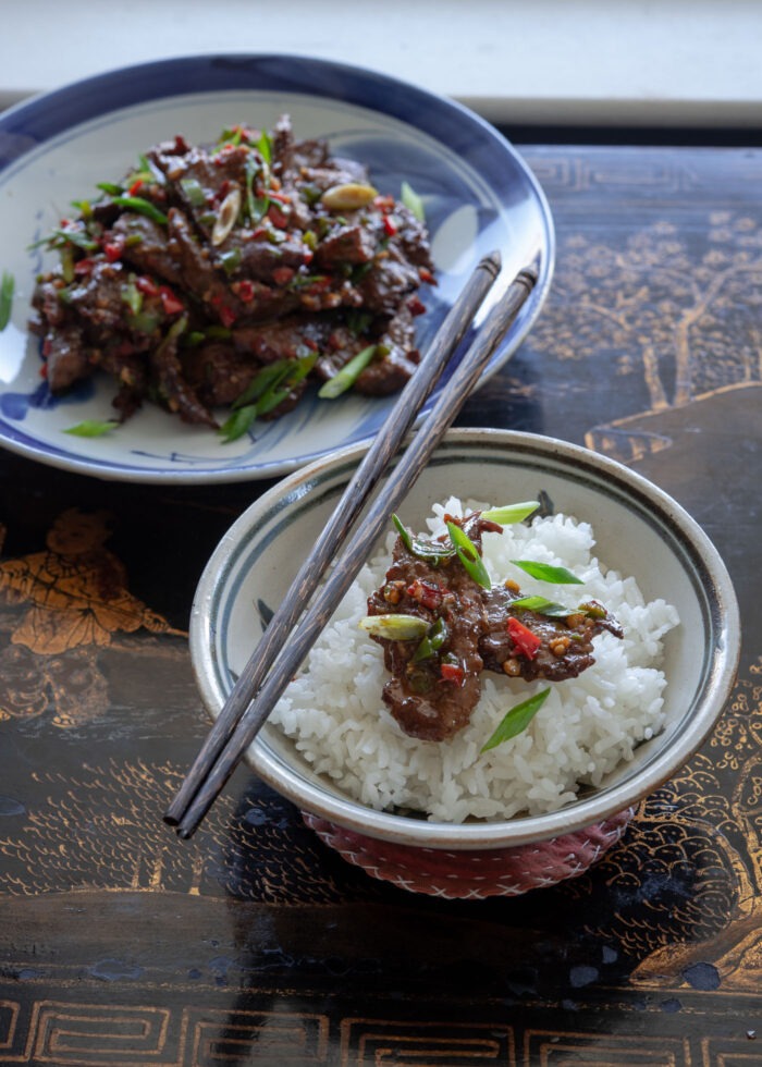 A bowl of rice is topped with a slice of cumin beef stir-fry and chopsticks.