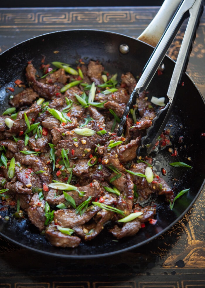 Beef stir-fry with cumin and chili is in a wok with kitchen tongs