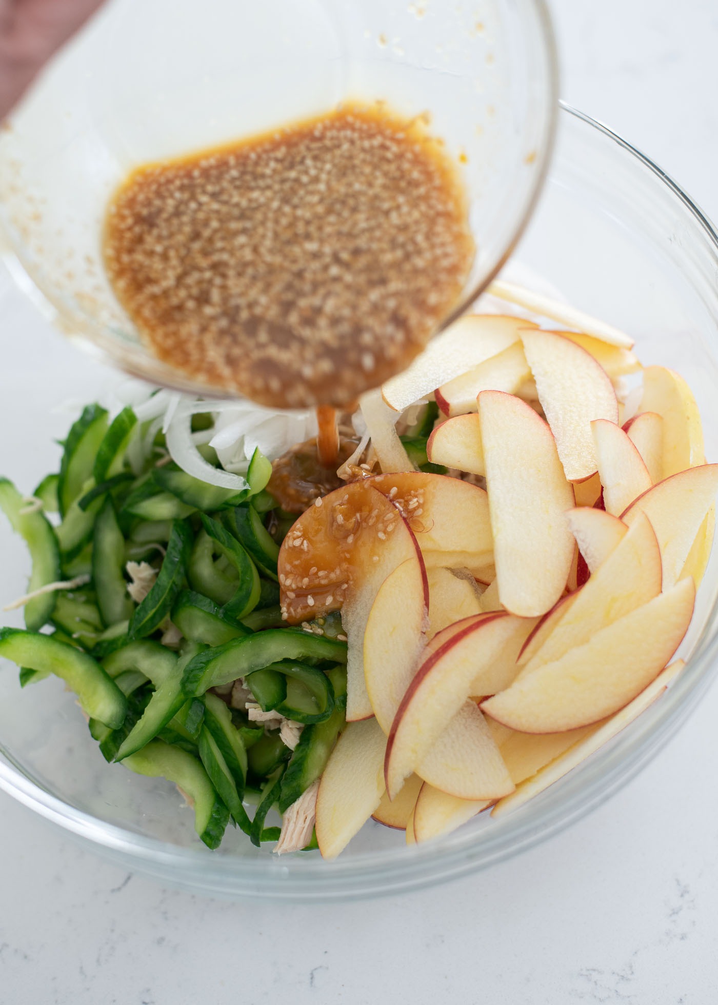 Sesame seed salad dressing is being poured on chicken, apple, and cucumber in a mixing bowl.