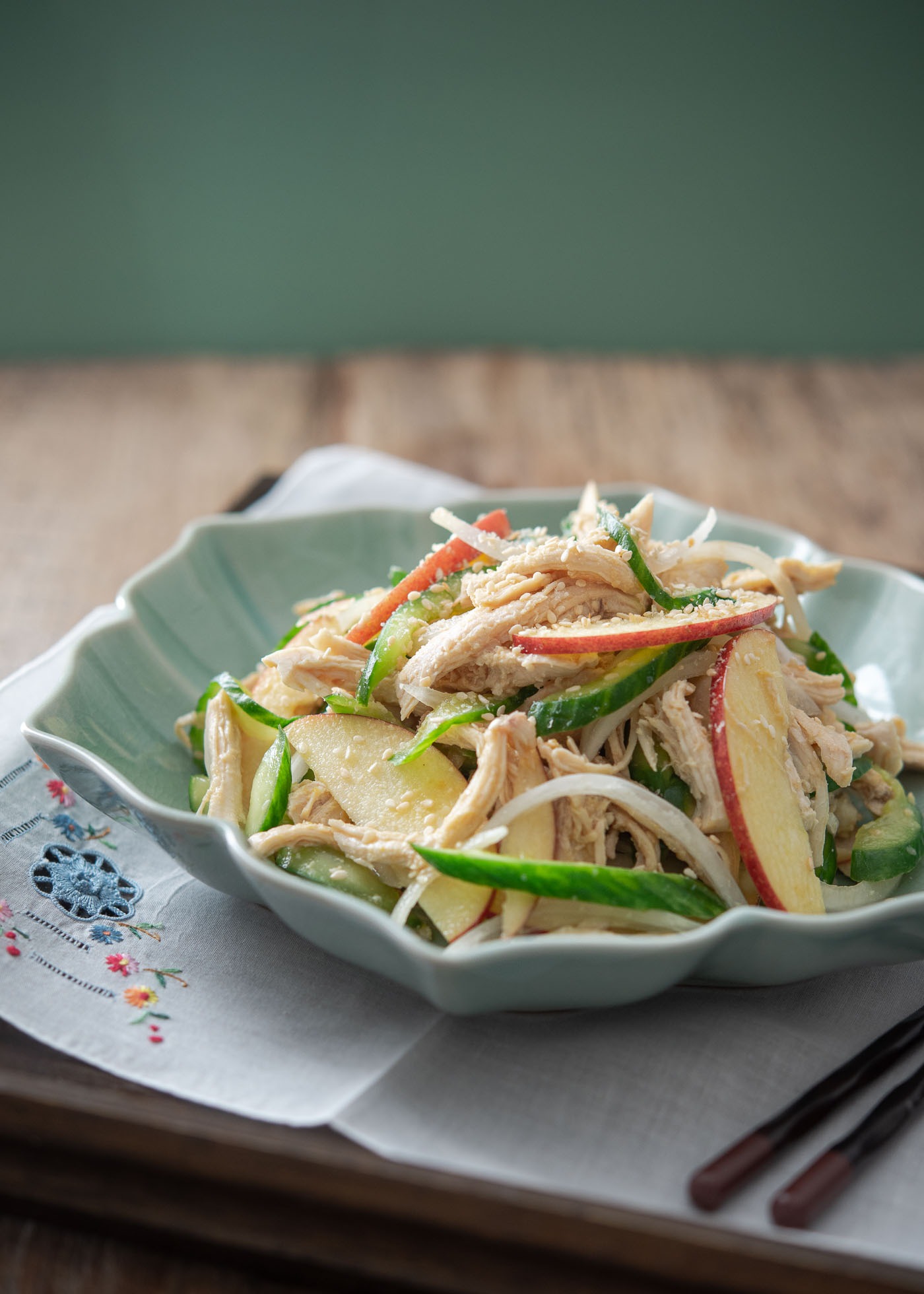 A teal colored Korean bowl is filled with chicken, cucumber, and apple salad on a tray lined with white napkin.