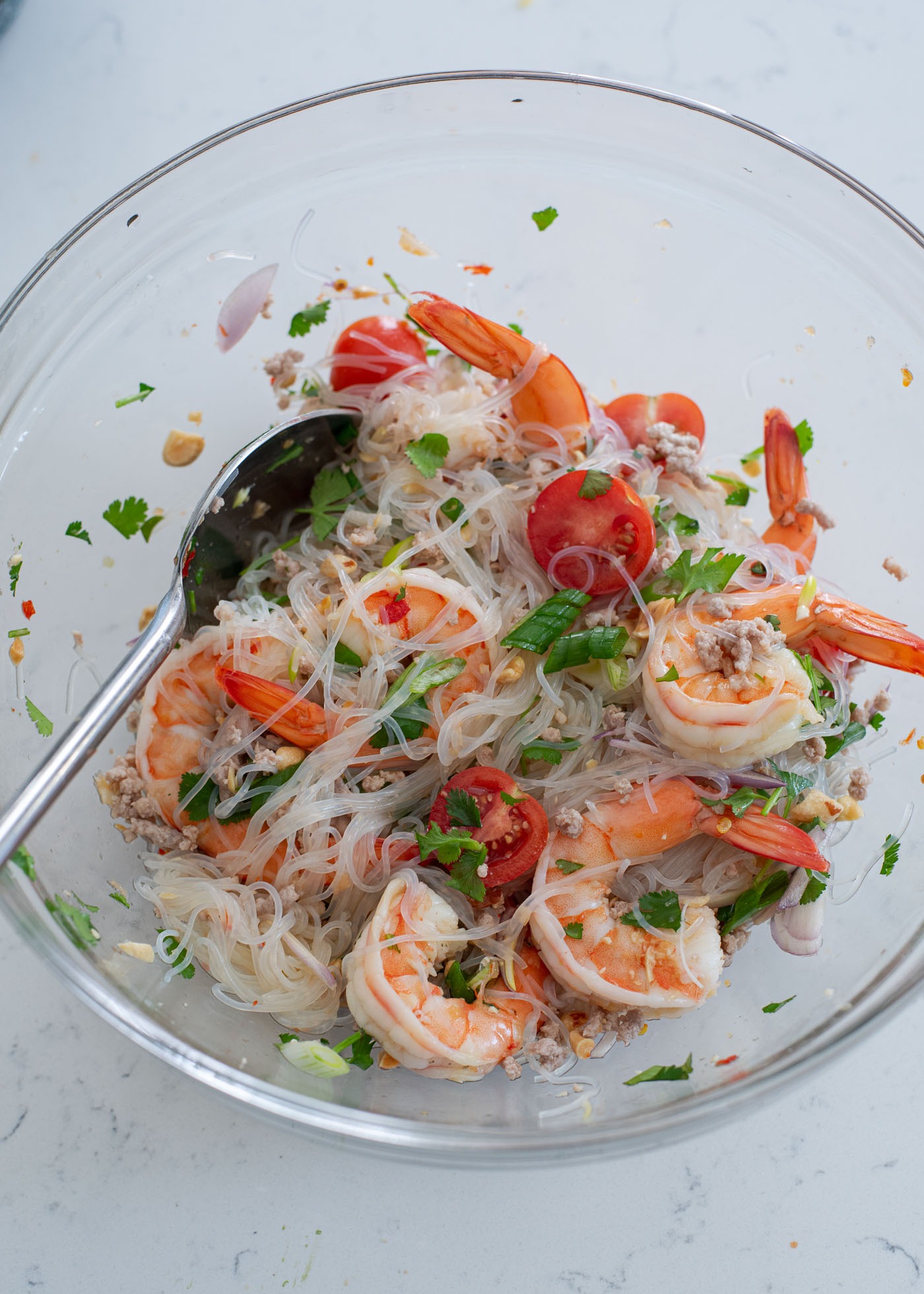A spoon is tossing shrimp, tomato, glass noodles with a salad dressing in a mixing bowl