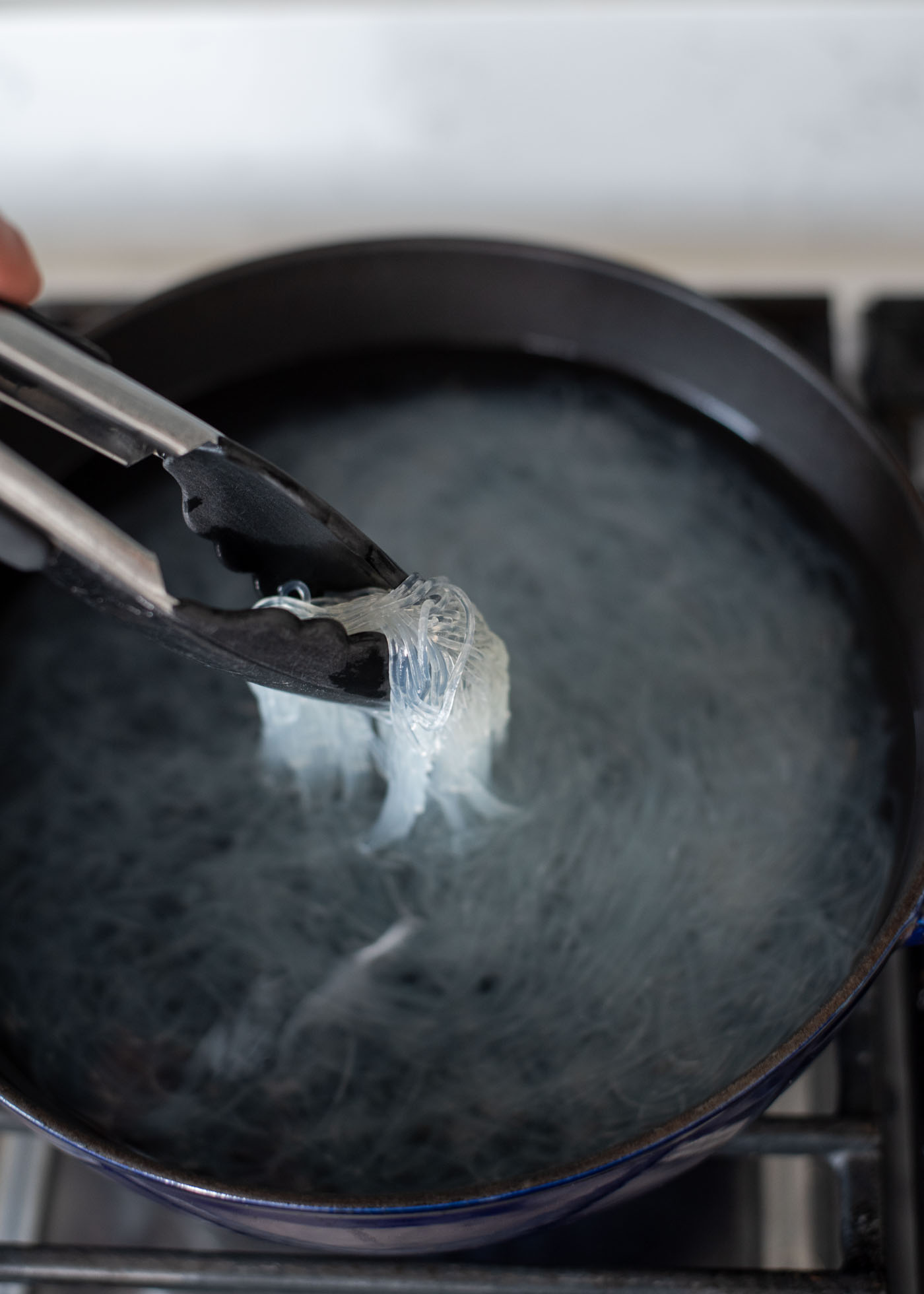 Kitchen tongs are picking up cooked white glass noodles from the simmering water.