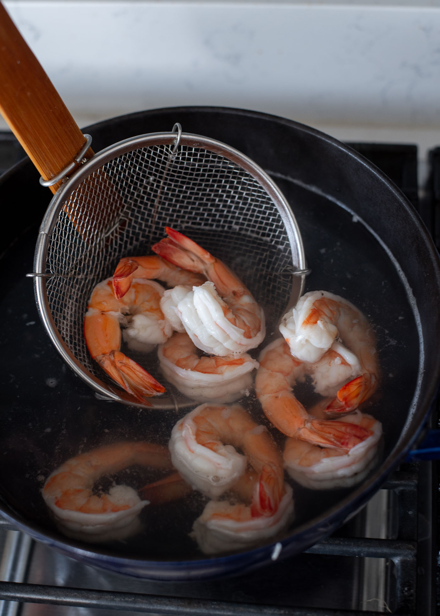 A wired skimmer is picking up cooked shrimp from the simmering water in a pot.