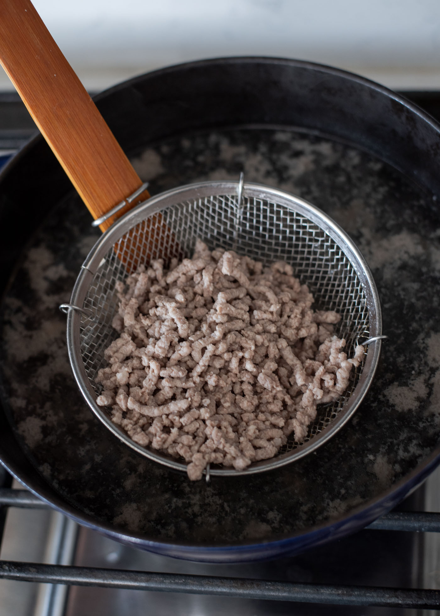 A wired skimmer is collecting cooked minced pork from the simmering water in a pot.