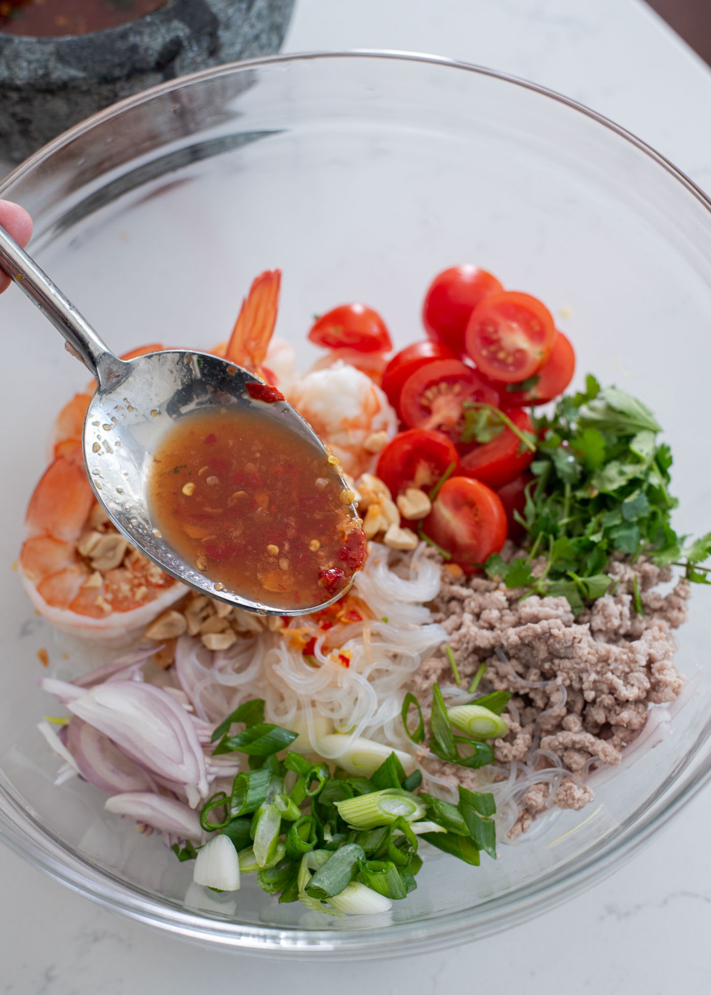 Thai chili and fish sauce dressing is spooned over glass noodle salad ingredients in a mixing bowl.