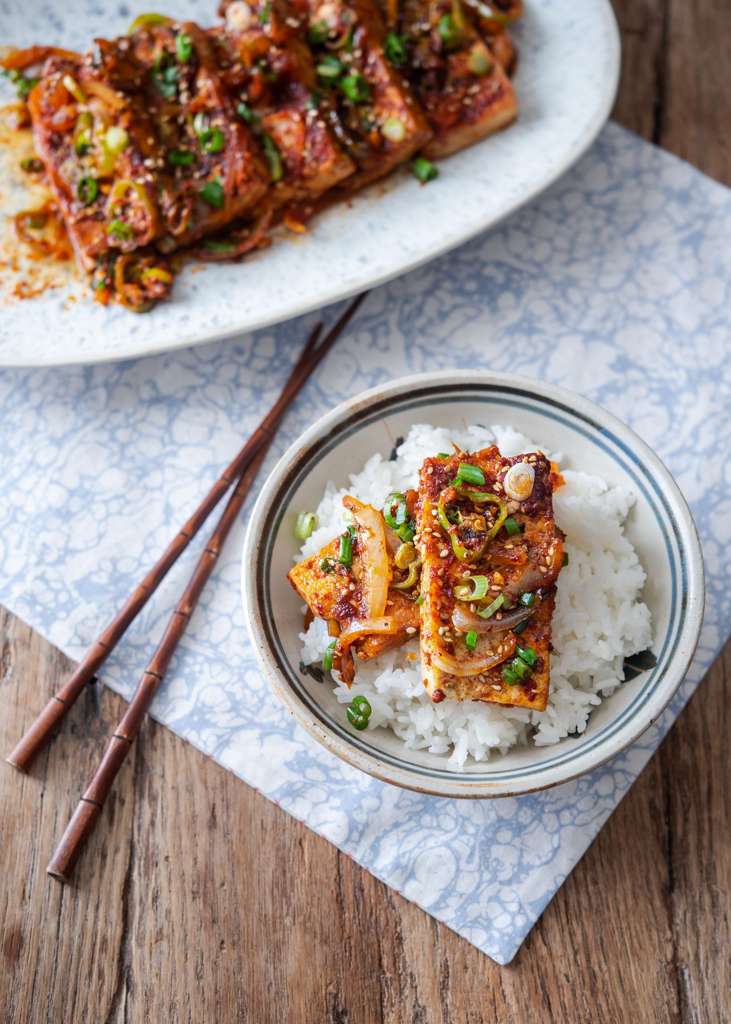 2 spicy tofu slices are placed on top of a bowl of rice next to chopsticks on a napkin.