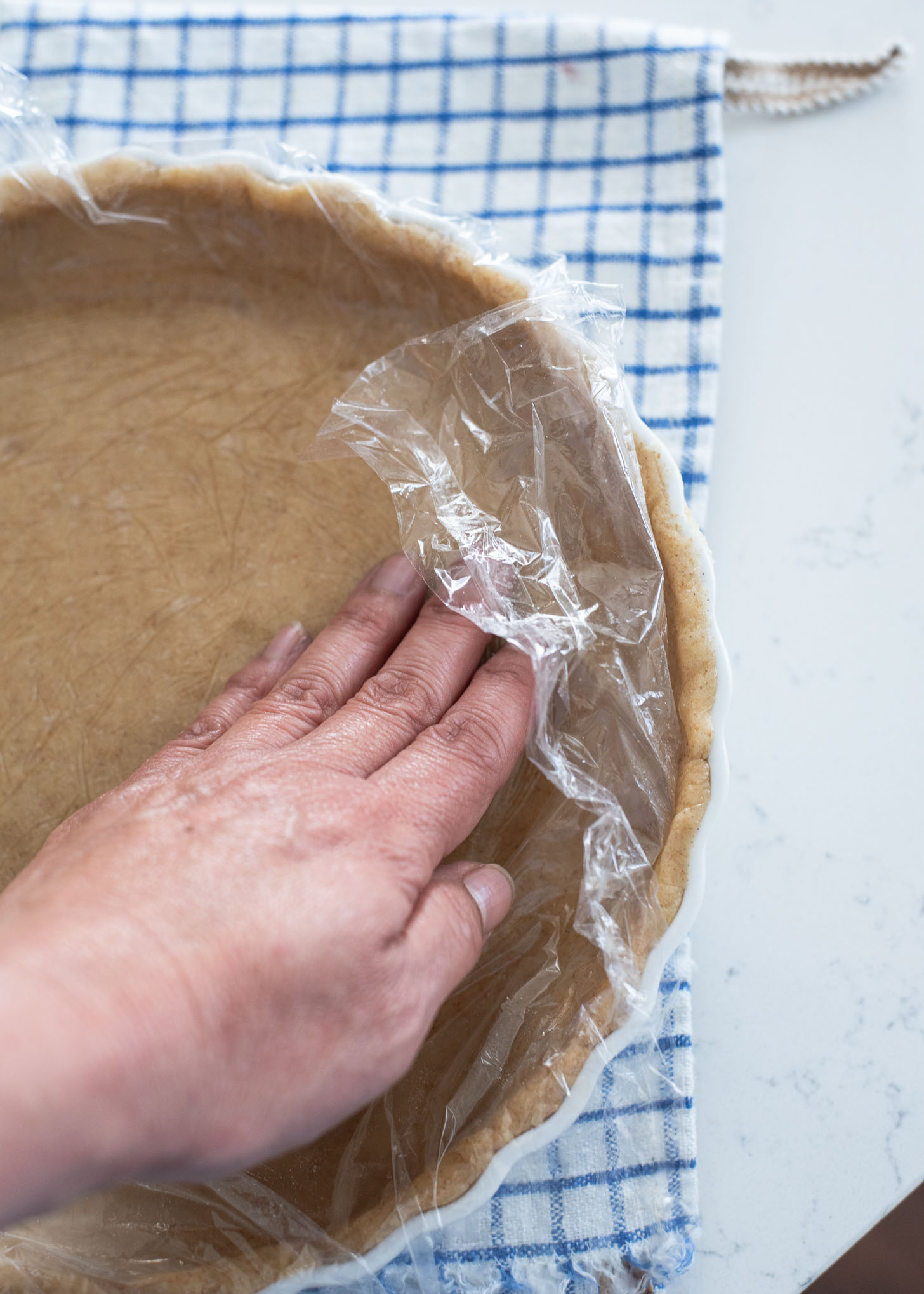 A pie dough covered with a plastic wrap is pressed to a pie dish by a hand.