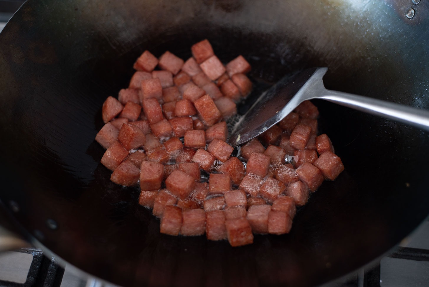 Diced Spam pieces are frying with oil in a wok.