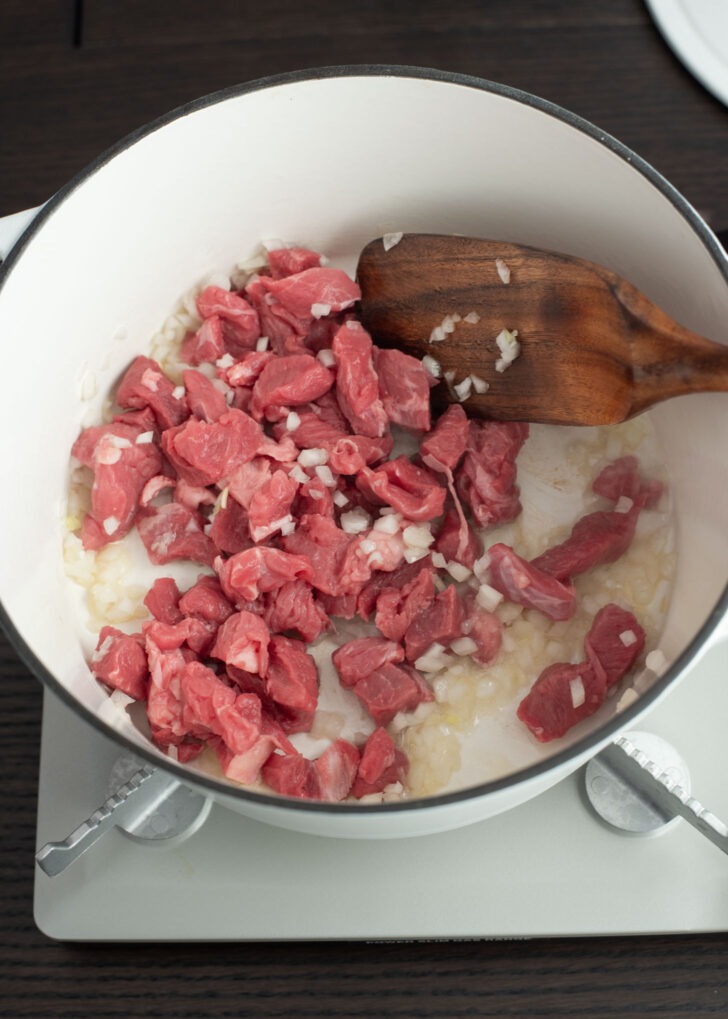 Beef pieces are added with chopped onion in a pot to cook.