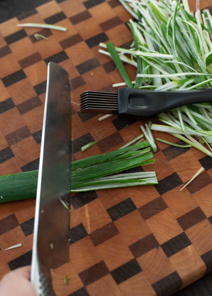 A knife is cutting off the portion of green onion what has been shredded.