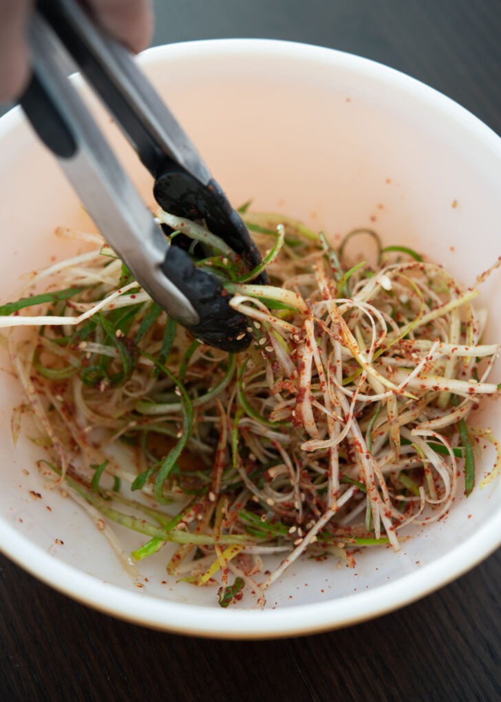 Korean green onion is being tossed with salad seasoning using kitchen tongs.