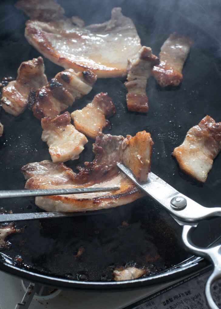 A pair of scissor is cutting of pork belly into bite size pieces.
