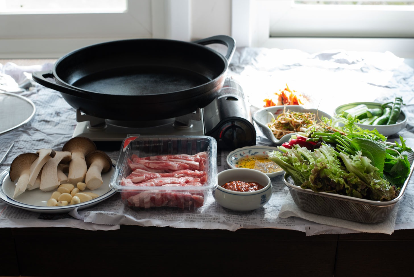 A large grill pan is placed over portable burner along with other dishes on the table for indoor BBQ.