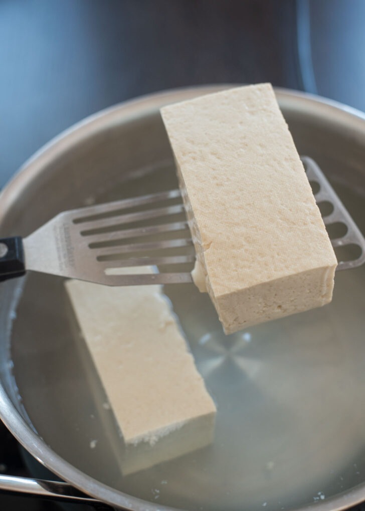 A spatula is lifting a block of tofu from the simmering water.
