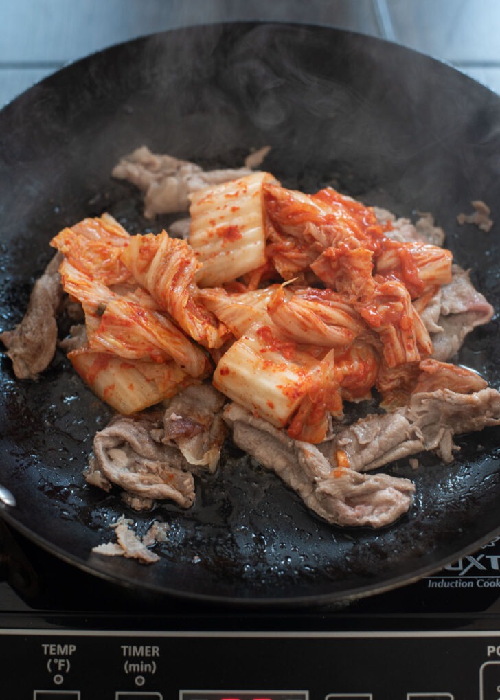 Sour kimchi pieces are added to cooked pork in a skillet.