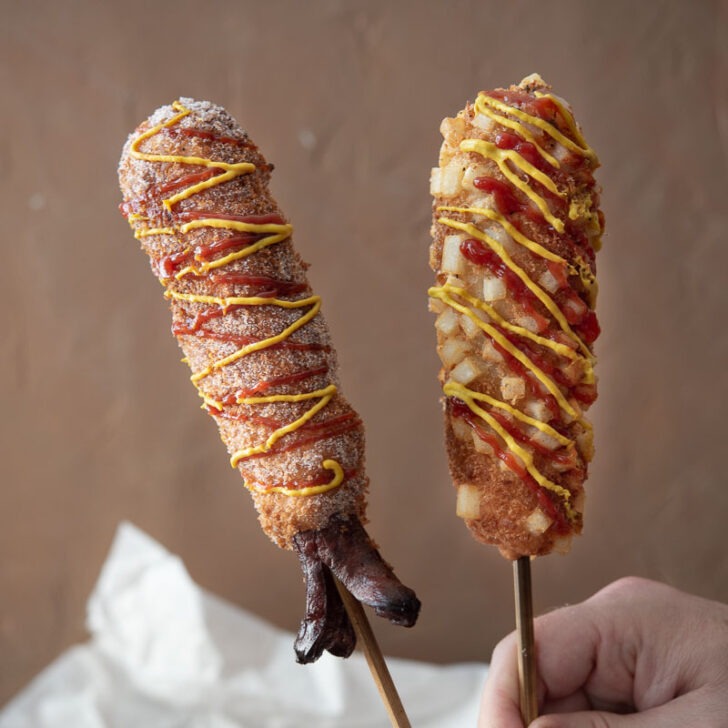 2 different types of Korean corn dogs that are garnished with condiments are being held upright.