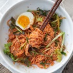 Bibim guksu Korean cold kimchi noodles lifted with chopsticks from a bowl topped with egg and vegetables