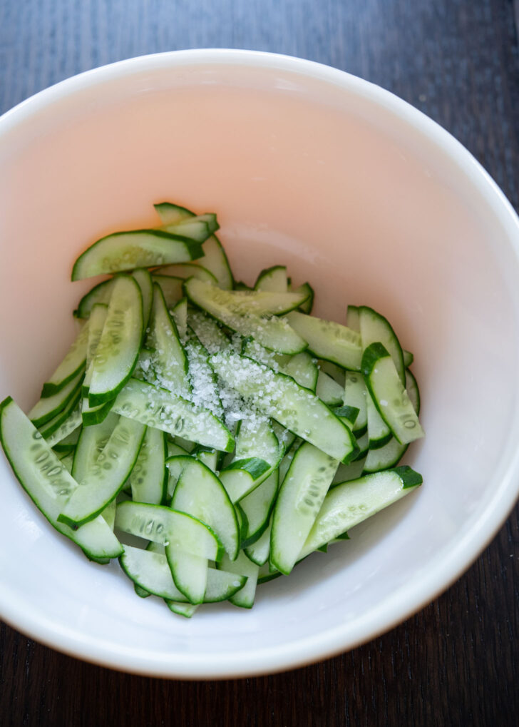 Salt is sprinkled over sliced cucumber.