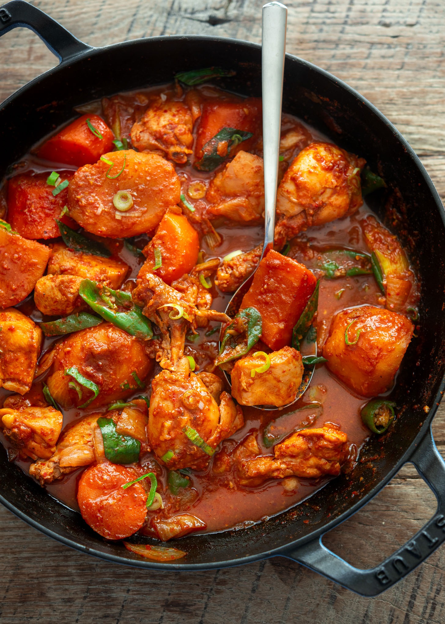 A serving spoon scooping out spicy chicken stew with carrot and potato from a pan.