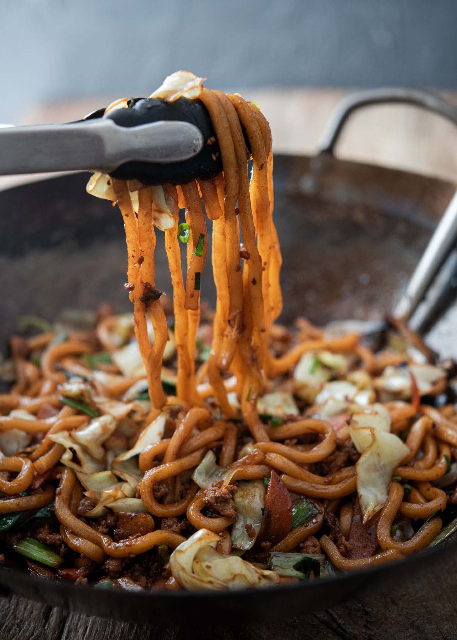 Kitchen tongs holding up yaki udon noodles from a wok.