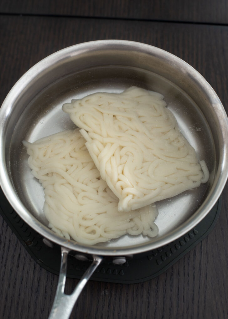 Frozen udon noodles soaking in a pot of hot water.