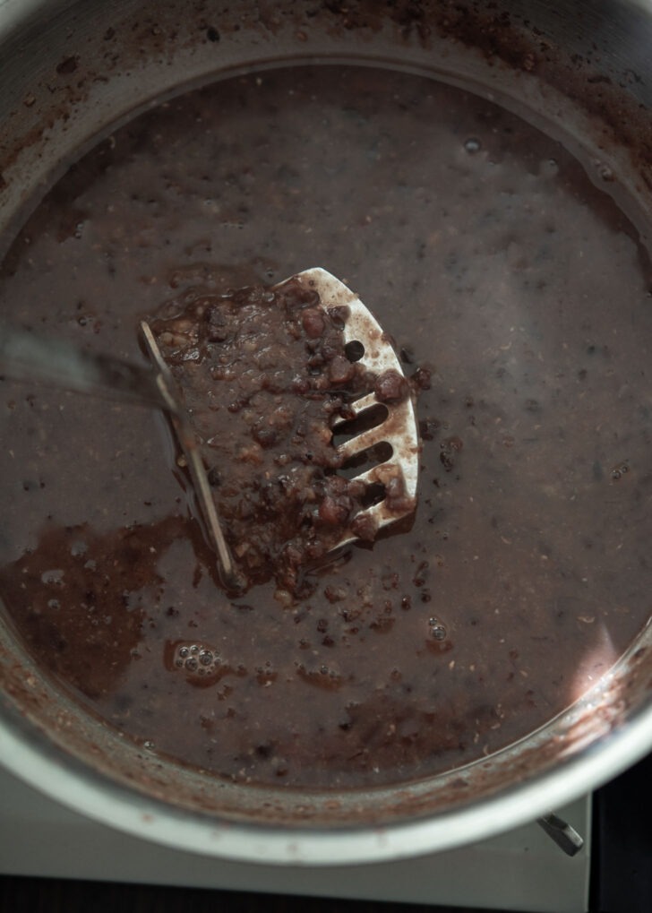 A potato masher used mashing soft red beans in a pot.