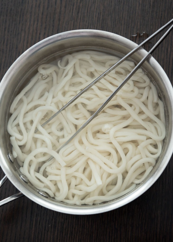Kitchen tongs separating softened udon noodles.