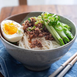 A bowl of noodles with ground beef sauce, cucumber, and hard boiled egg.