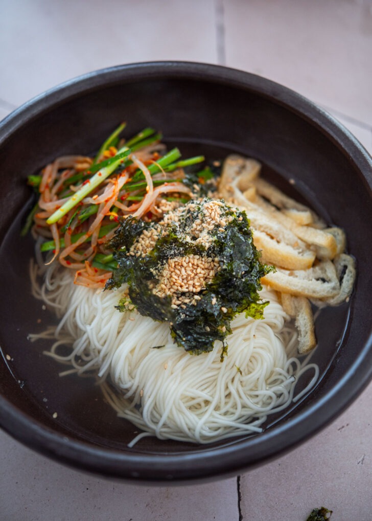 Pouring hot anchovy broth into a bowl of somyeon noodles with vegetable toppings.