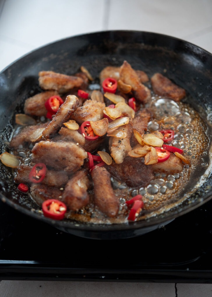 Crispy pork and sliced red chili added to thickened garlic glaze in a skillet
