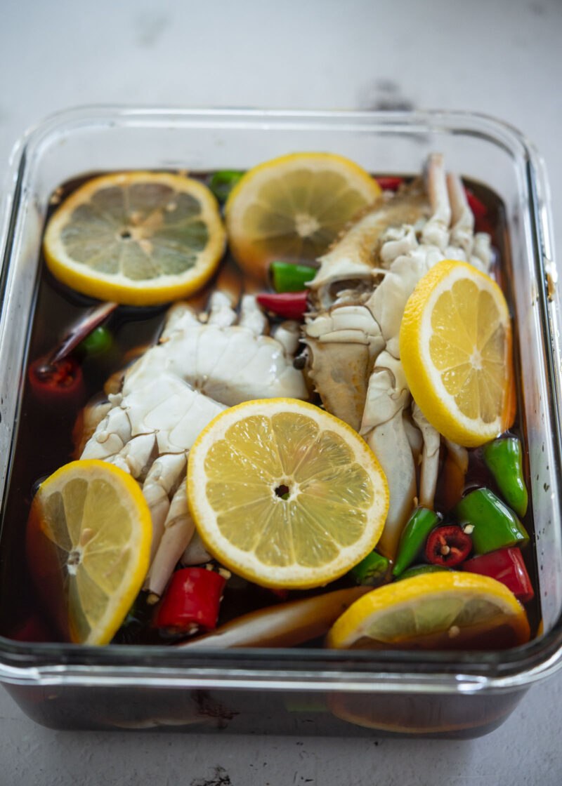 Crabs covered in soy marinade with lemon slices and chilies in a glass container.