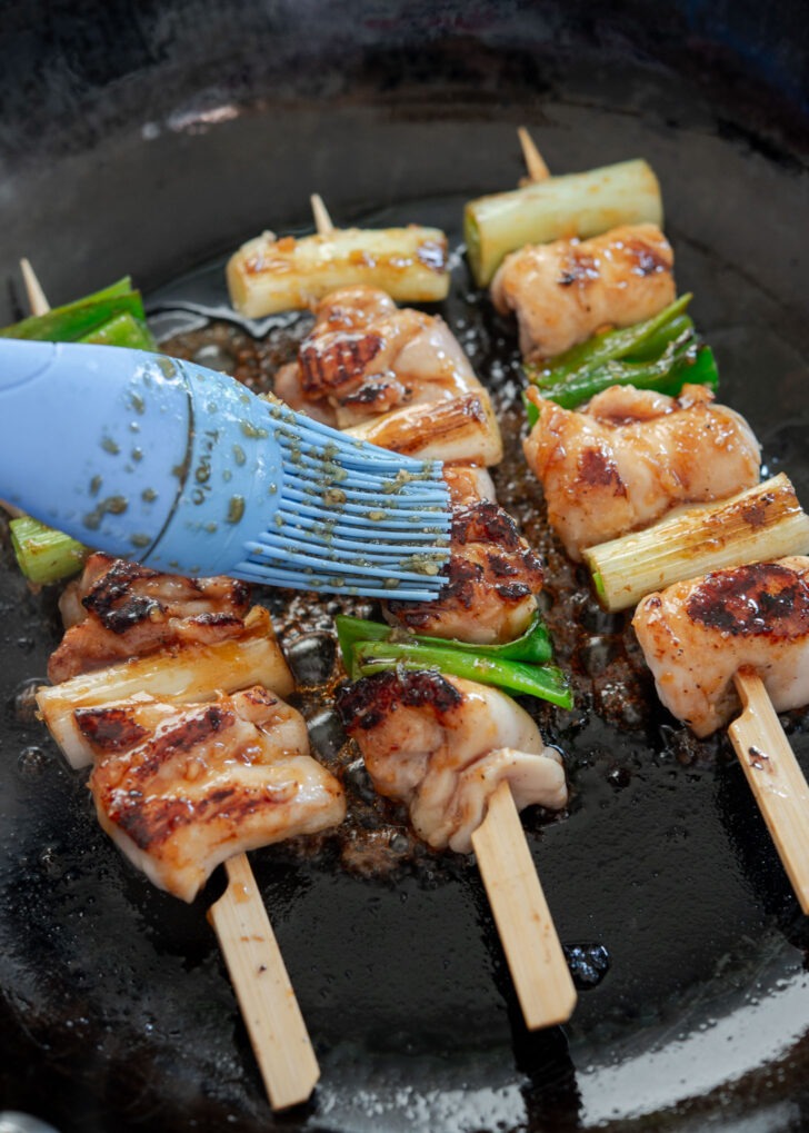 Brushing soy-garlic glaze onto dakkochi chicken skewers in a skillet
