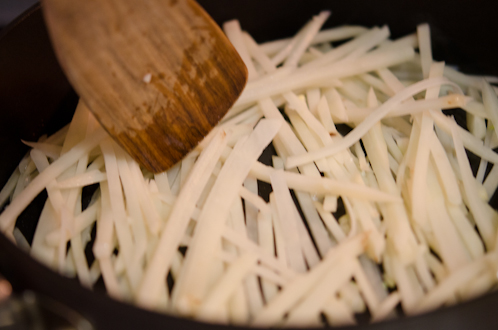 Potato matchstick slices are being tossed with a wooden spatula.