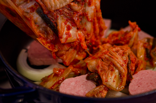 Sour kimchi is being added to Spam and onion mixture in a pot.