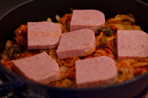 More Spam slices are added on top of kimchi in a pot.