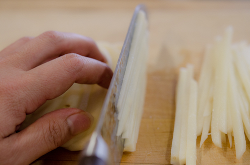 Thinly sliced potatoes are being cut into thin matchsticks with a knife.